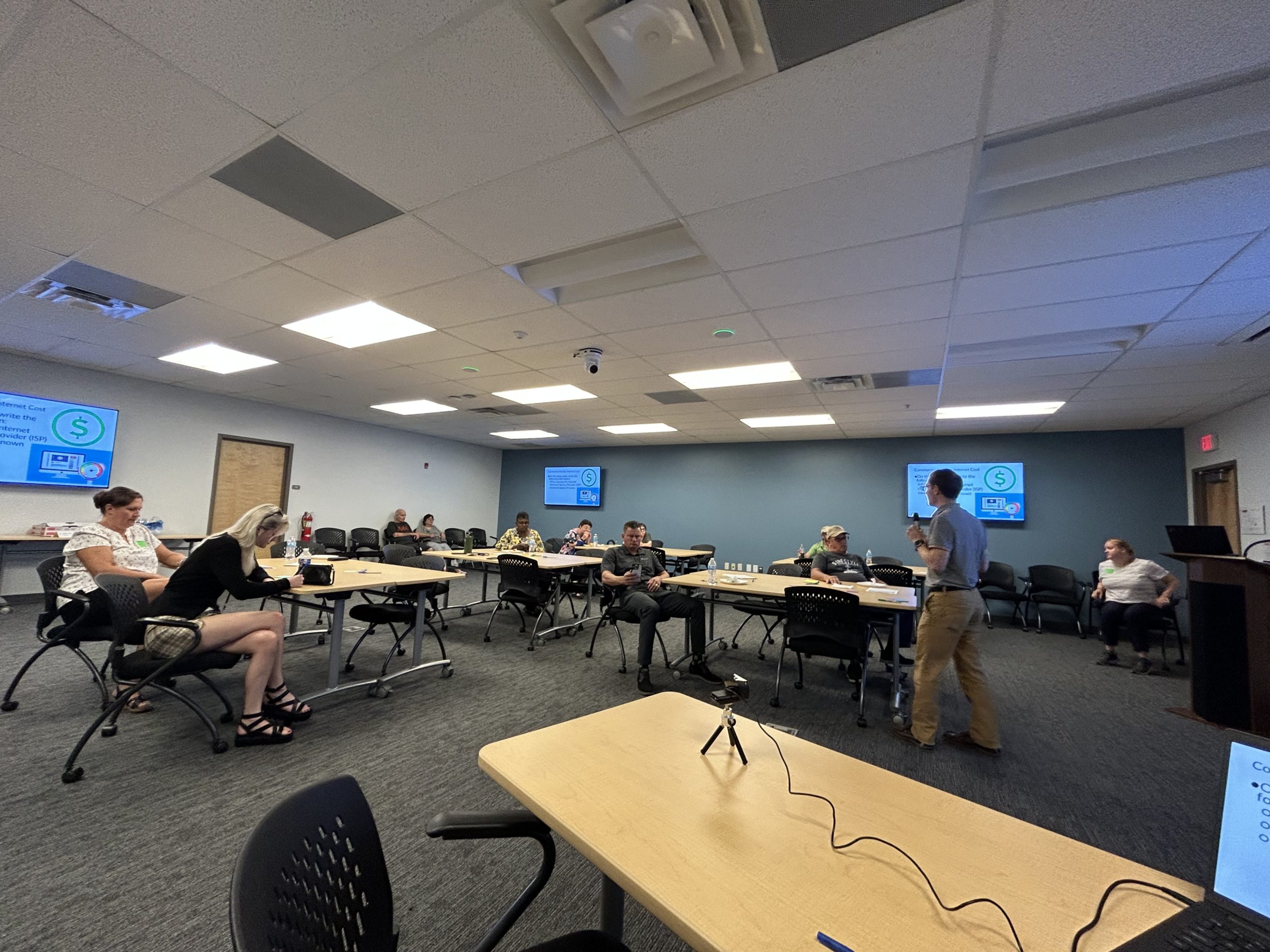 Sean Fox presenting in gray shirt and khakis to a room of 8 individuals sitting at tables